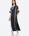 A woman wears the Sea New York Juliet Polo Maxi Dress— a loose, short-sleeved style with vertical color-block stripes in navy blue, green, beige, and gold—paired with classic white oxford shoes.