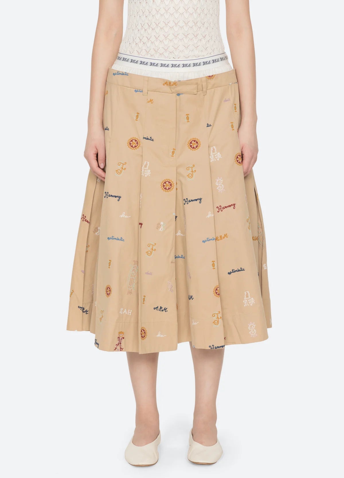 Wearing the Sea New York Juliet Skirt—a beige, knee-length embroidered skirt with colorful patterns and text—paired with a white textured top and white shoes. The photo is cropped just below the shoulders.