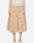 Wearing the Sea New York Juliet Skirt—a beige, knee-length embroidered skirt with colorful patterns and text—paired with a white textured top and white shoes. The photo is cropped just below the shoulders.
