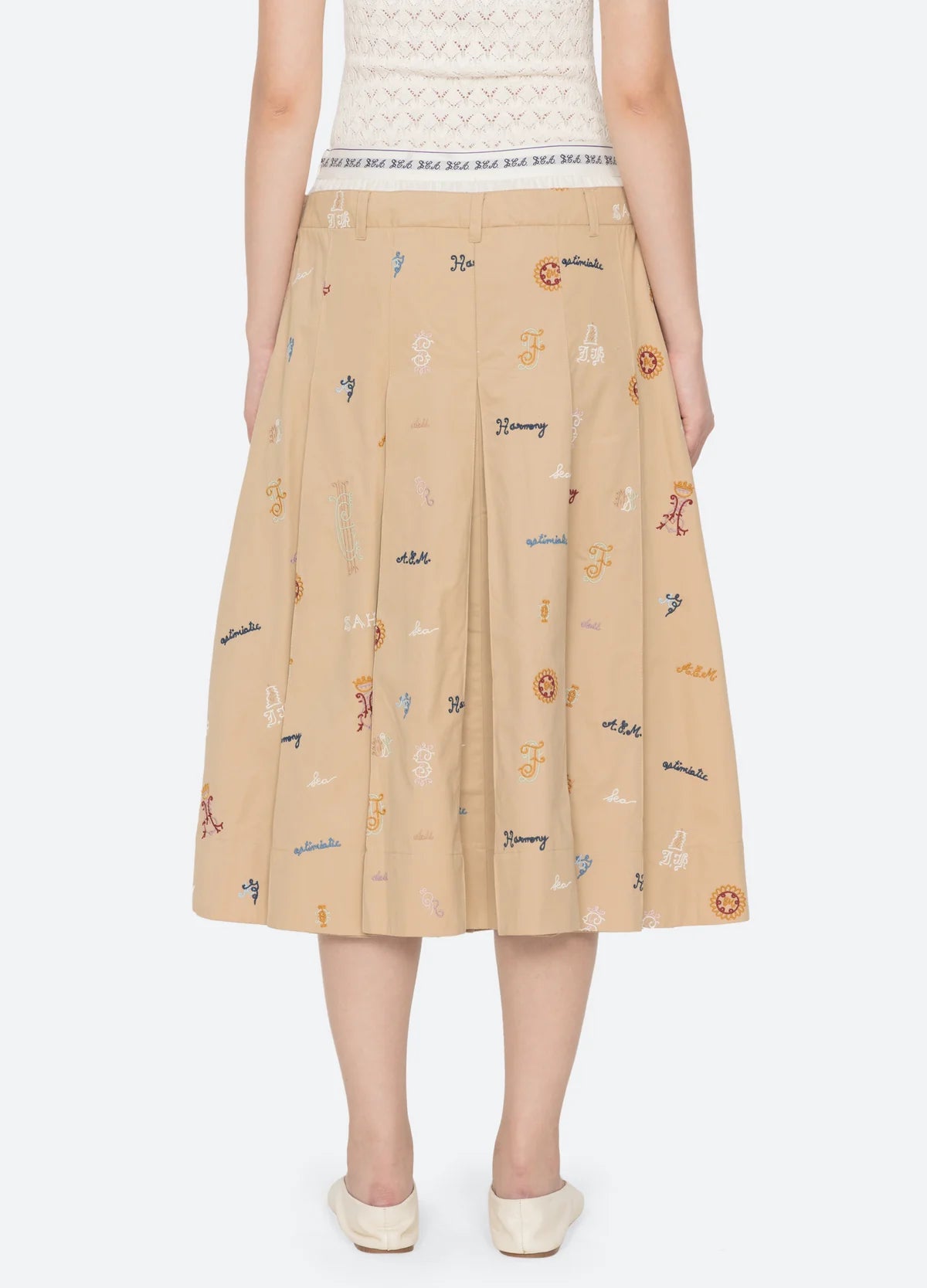 A person stands facing away, wearing the Sea New York Juliet Skirt—a beige pleated skirt with an athletic waistband and colorful embroidered motifs—paired with a white top and beige flats.