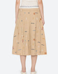 A person stands facing away, wearing the Sea New York Juliet Skirt—a beige pleated skirt with an athletic waistband and colorful embroidered motifs—paired with a white top and beige flats.