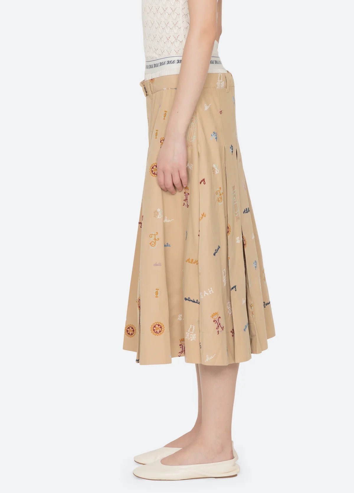 A person in profile wears the Sea New York Juliet Skirt—a beige pleated skirt with colorful embroidery and an athletic waistband—with a white sleeveless top and white flats, set against a plain light background.