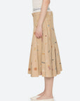 A person in profile wears the Sea New York Juliet Skirt—a beige pleated skirt with colorful embroidery and an athletic waistband—with a white sleeveless top and white flats, set against a plain light background.