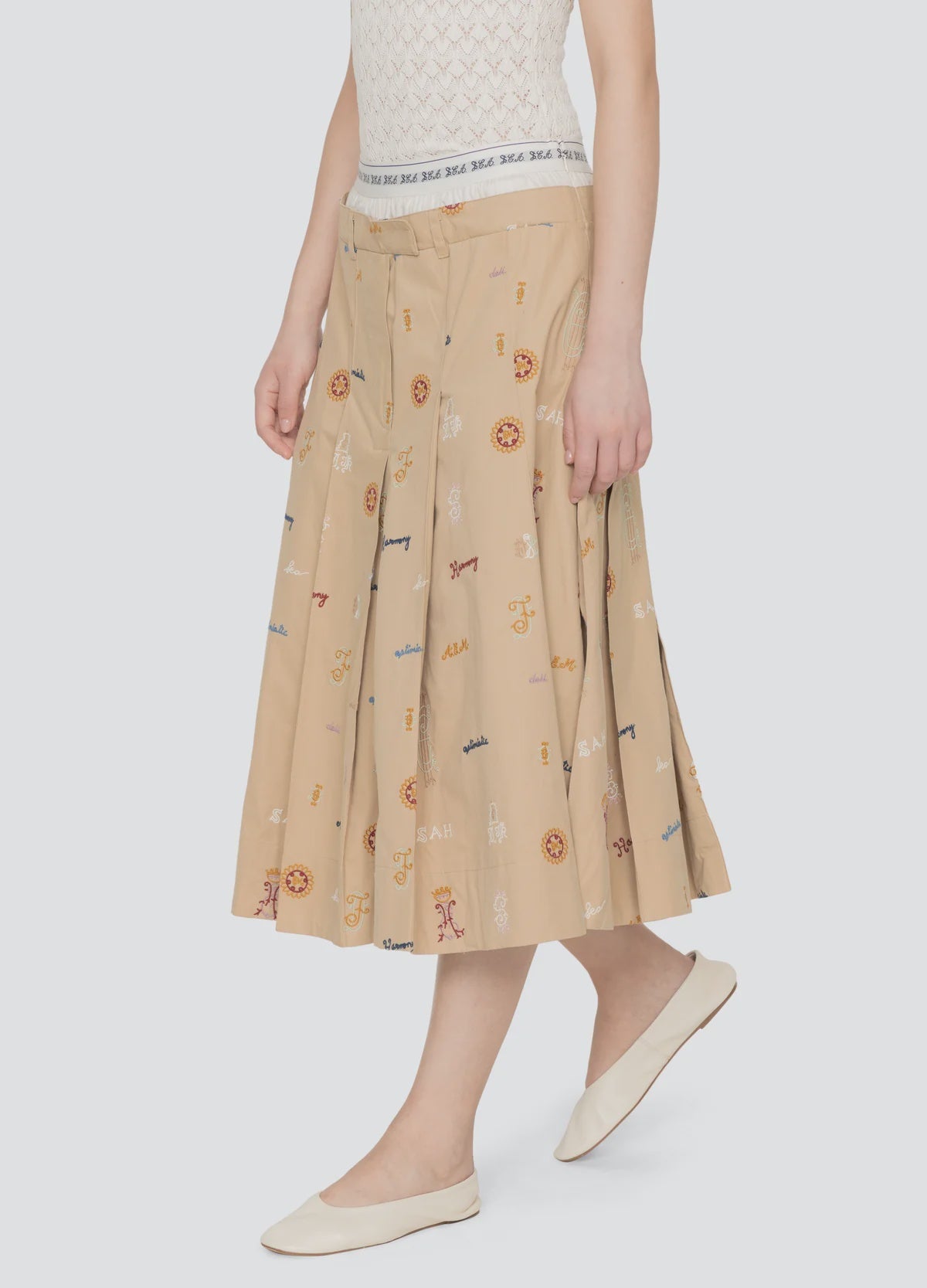 A person wears the Sea New York Juliet Skirt—a beige pleated embroidered skirt with colorful symbols and an athletic waistband—paired with a white lace top and white ballet flats, standing against a plain light background.
