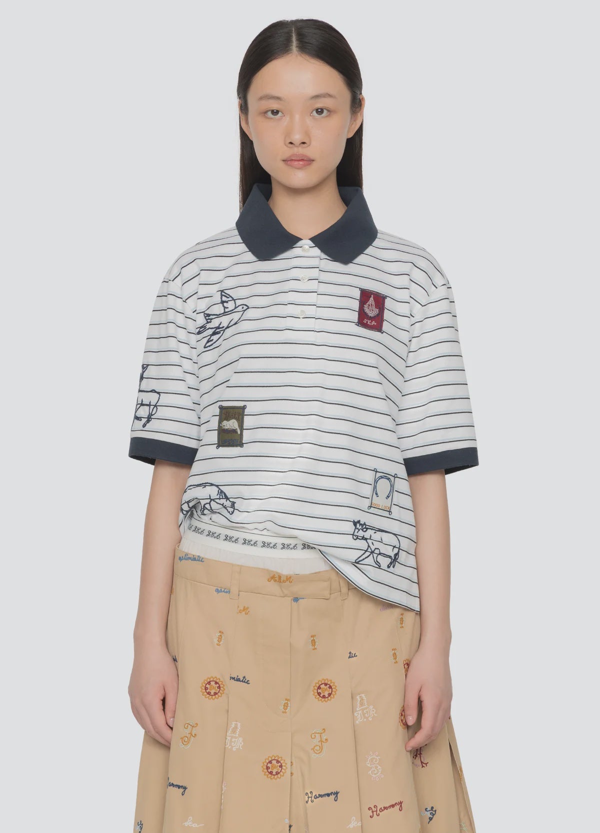 A young woman wears the Winslow Craft Polo Shirt by Sea New York, a relaxed-fit, short-sleeved top with embroidered animals and patches, paired with a beige skirt with colorful patterns. She stands against a plain light background.