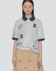 A young woman wears the Winslow Craft Polo Shirt by Sea New York, a relaxed-fit, short-sleeved top with embroidered animals and patches, paired with a beige skirt with colorful patterns. She stands against a plain light background.