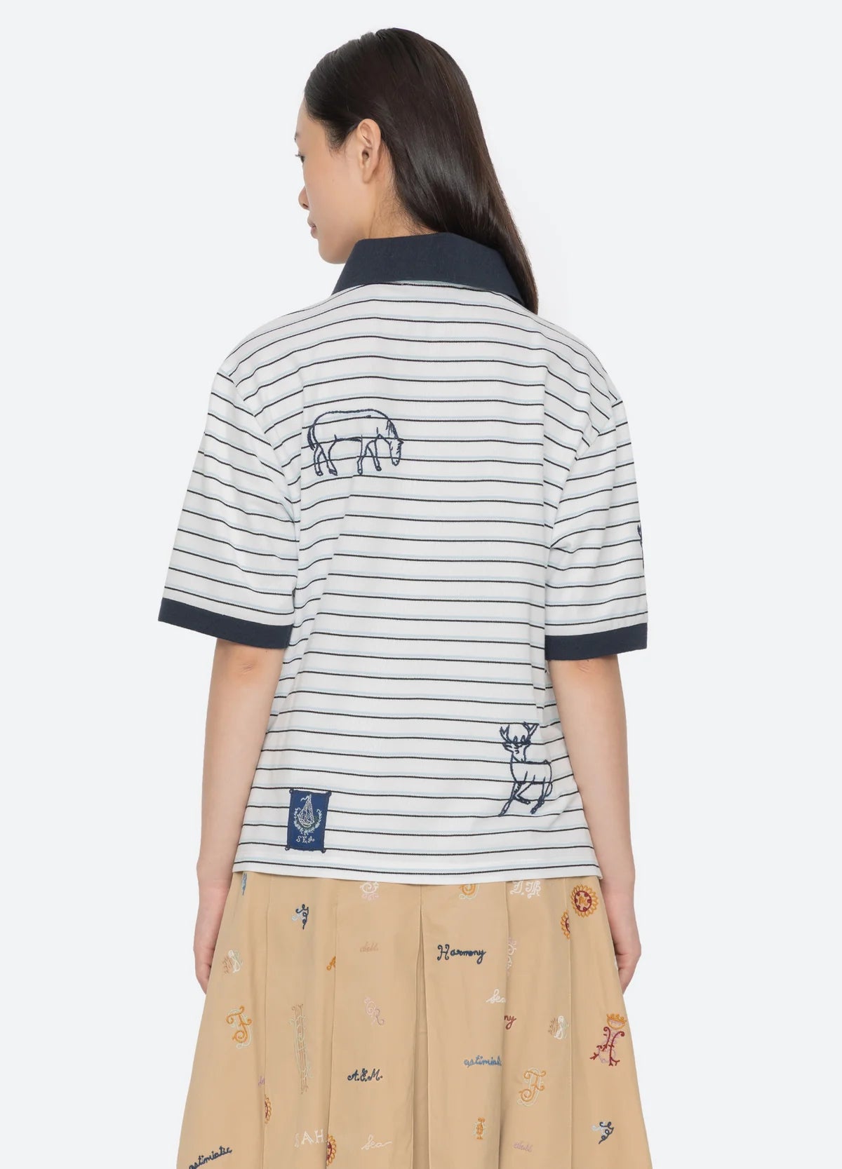 A woman with long dark hair wears the Sea New York Winslow Craft Polo Shirt, a relaxed-fit white and navy striped polo with embroidered animals on the back, paired with a beige skirt featuring colorful embroidery.