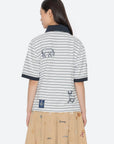 A woman with long dark hair wears the Sea New York Winslow Craft Polo Shirt, a relaxed-fit white and navy striped polo with embroidered animals on the back, paired with a beige skirt featuring colorful embroidery.
