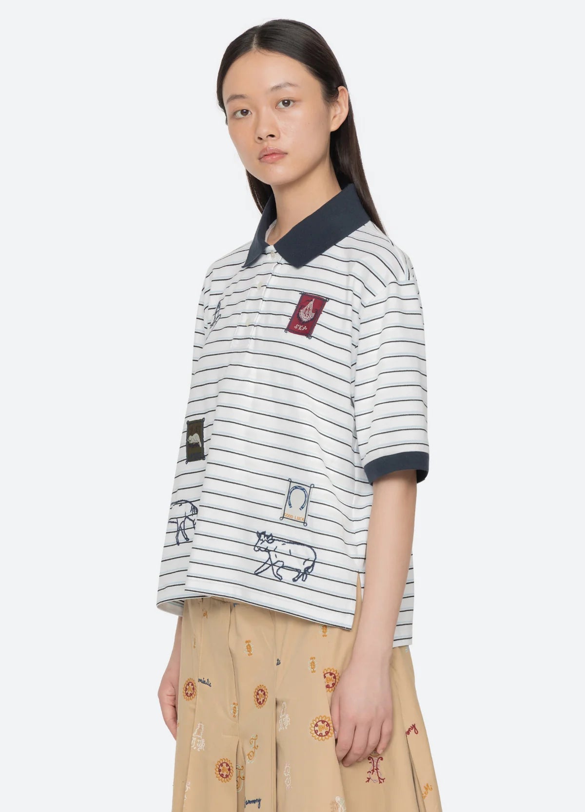 A young woman with straight dark hair wears the Winslow Craft Polo Shirt by Sea New York, a relaxed-fit white top with black stripes and embroidered animals, paired with a tan printed skirt. She stands against a plain light background, looking slightly to the side.