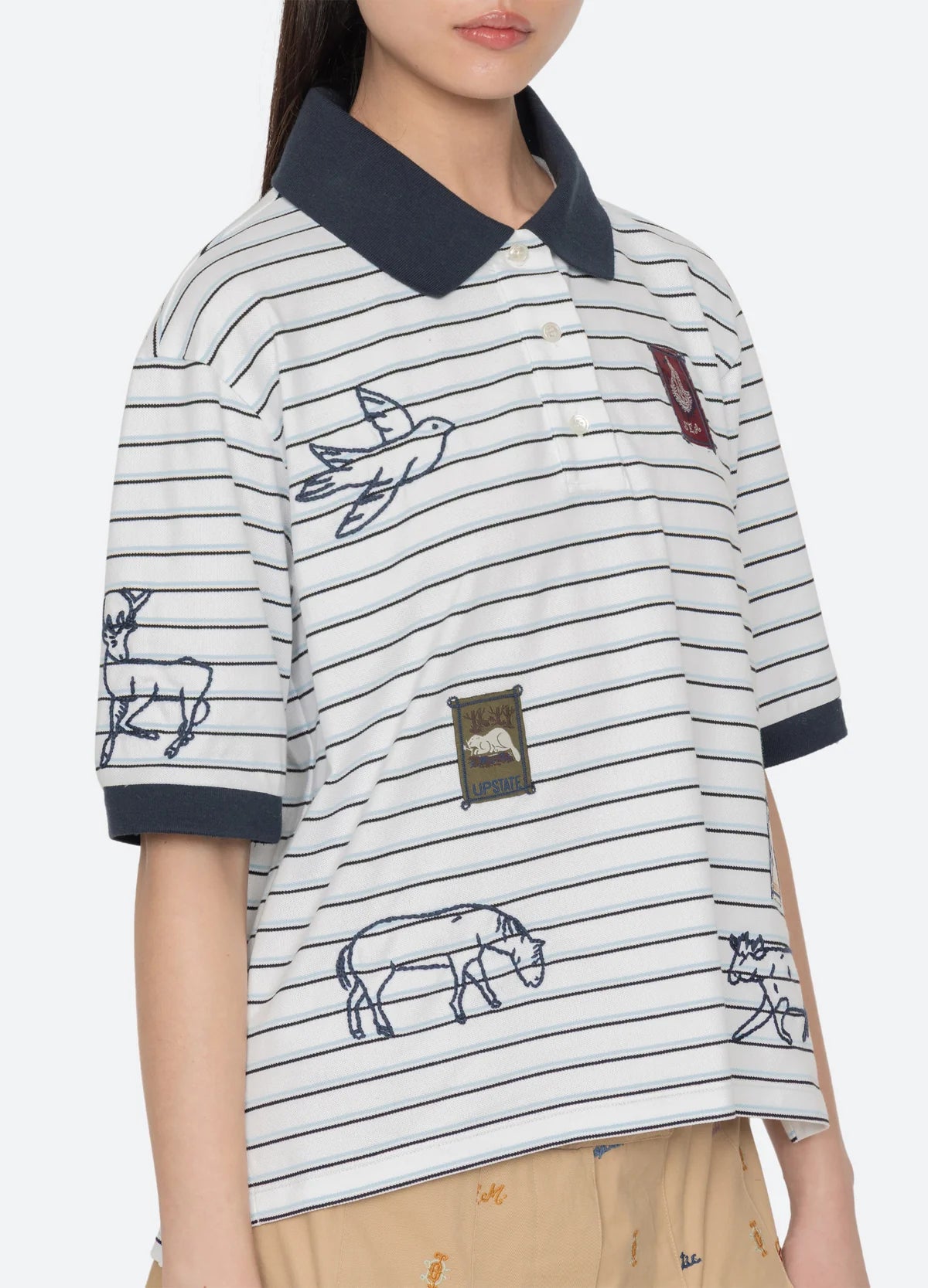 Wearing the Sea New York Winslow Craft Polo Shirt, this person sports a relaxed fit, short sleeves, striped design with embroidered animals, bird details, decorative patches, and dark collar and cuffs. Face is partially out of frame.