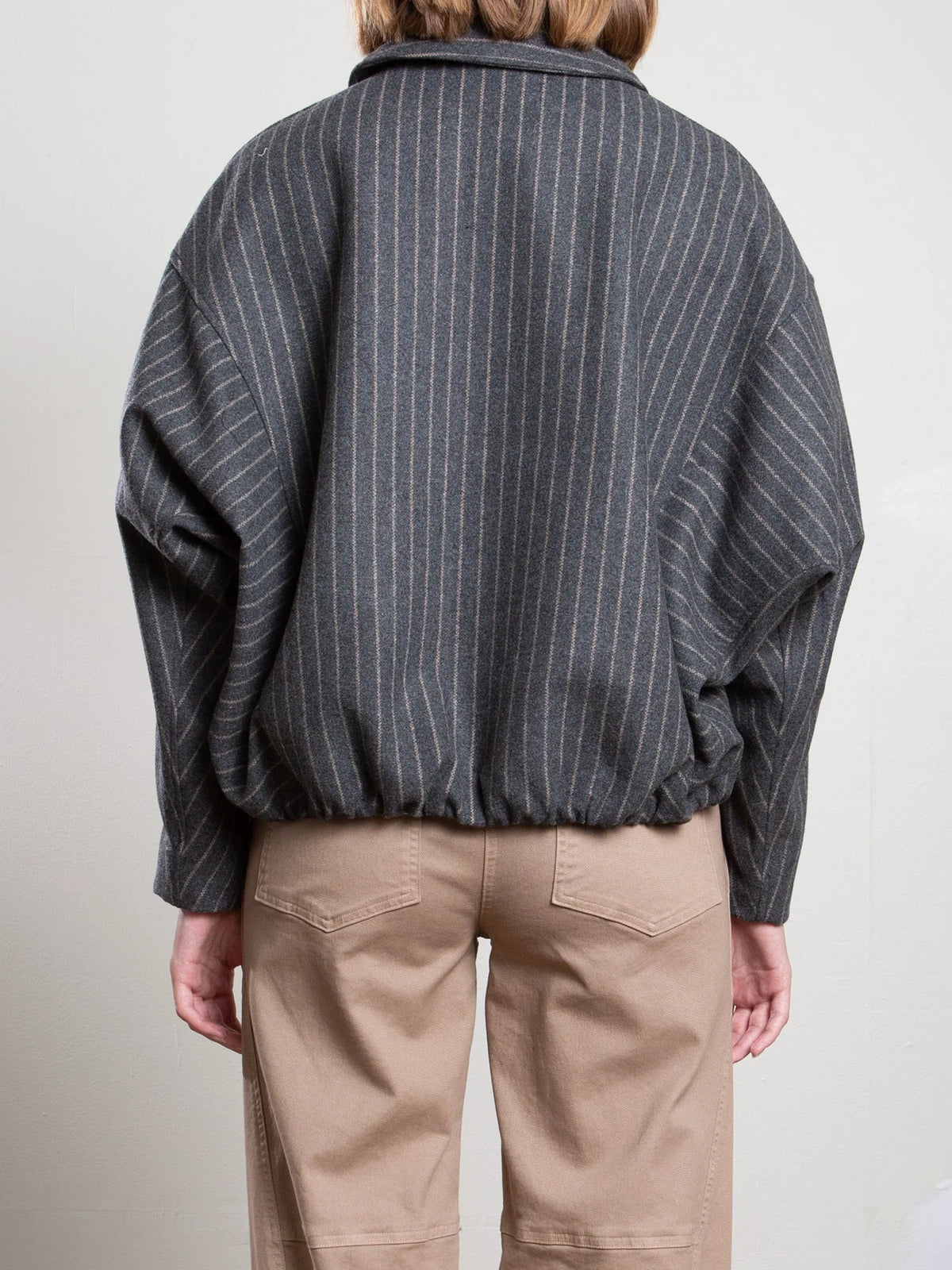 Pharaoh’s Andre Bomber Jacket in wool pinstripe, tan pants, back view; minimalist setting with plain wall.