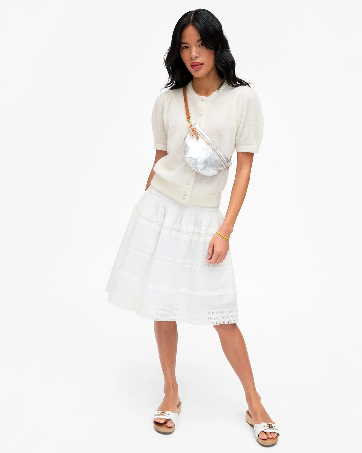 Woman wearing a white short-sleeve cardigan, white skirt, white slide sandals, and the Clare Vivier Petite Fanny S26 silver crossbody bag with adjustable strap, standing against a plain white background.