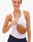 A woman in a white tank top and dark pants smiles while holding the Clare Vivier Petite Fanny S26—a small brown woven crossbody bag with an adjustable strap, green accent, and blue charm.