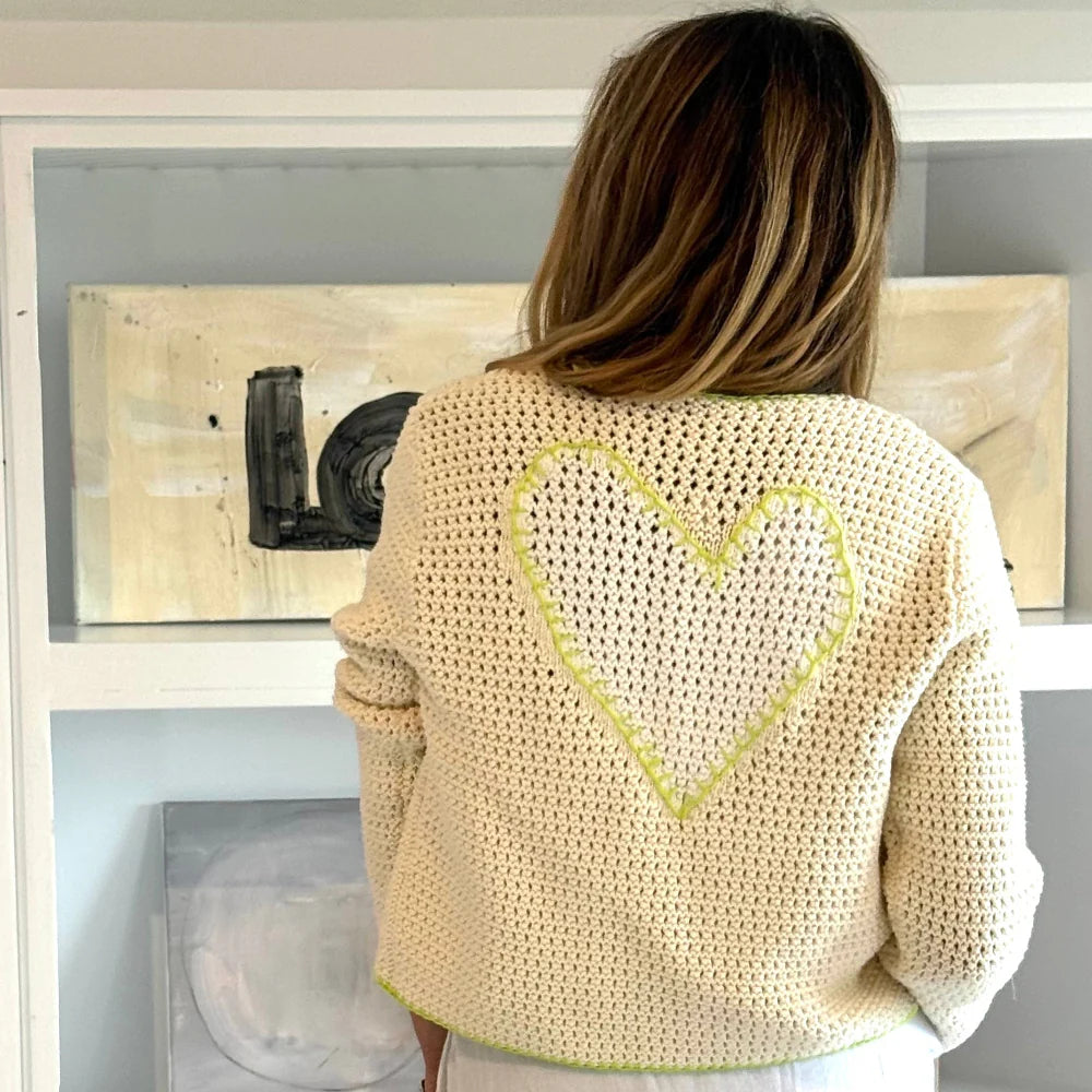 A person with long light brown hair faces away, wearing the Kerri Rosenthal Frankie Beau-Lero cotton crochet cardigan featuring a yellow-outlined heart on the back. Abstract art sits on white shelves behind them.