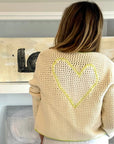 A person with long light brown hair faces away, wearing the Kerri Rosenthal Frankie Beau-Lero cotton crochet cardigan featuring a yellow-outlined heart on the back. Abstract art sits on white shelves behind them.