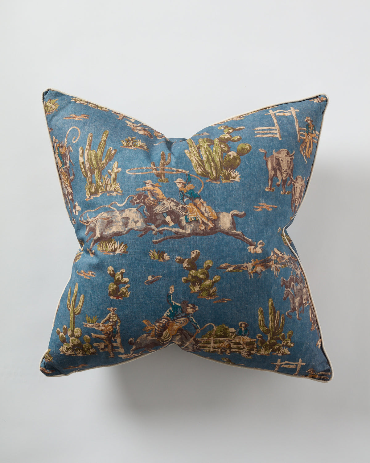 The Design Legacy Denim Cowboy Pillow 26x26 features a vintage Western print with cowboys, horses, cattle, cacti, and desert scenery on denim blue. Shown from above against a white surface.