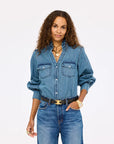 A woman with long curly hair wears the Kerri Rosenthal Puff Sleeve Shirt tucked into high-waisted jeans, accessorized with gold jewelry, a black belt, and a gold watch, standing against a plain white background.
