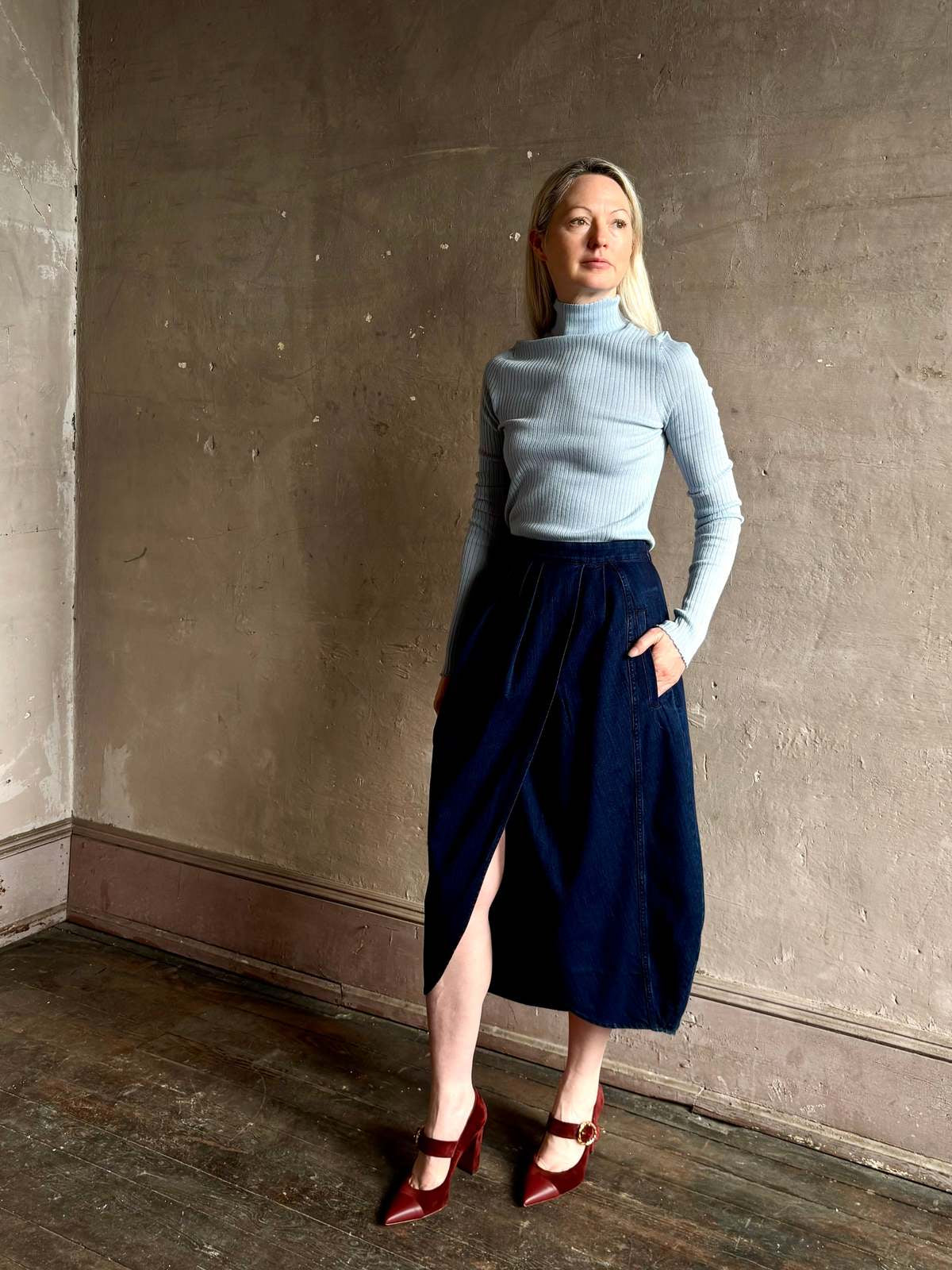 Rachel Comey Sobre Skirt in blue, styled with a turtleneck and red heels against a textured wall.