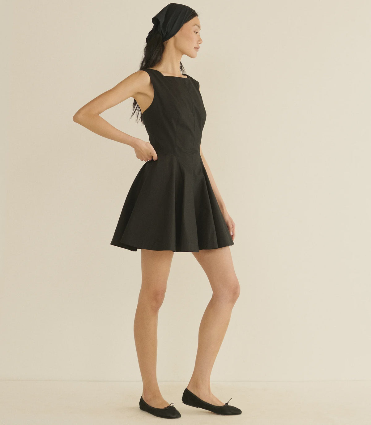 A woman stands sideways against a neutral background, wearing the DOEN Riviette Dress in Italian poplin with a flared mini skirt, paired with a black headscarf and black flats. Her hand rests on her waist as she looks forward.