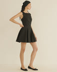 A woman stands sideways against a neutral background, wearing the DOEN Riviette Dress in Italian poplin with a flared mini skirt, paired with a black headscarf and black flats. Her hand rests on her waist as she looks forward.