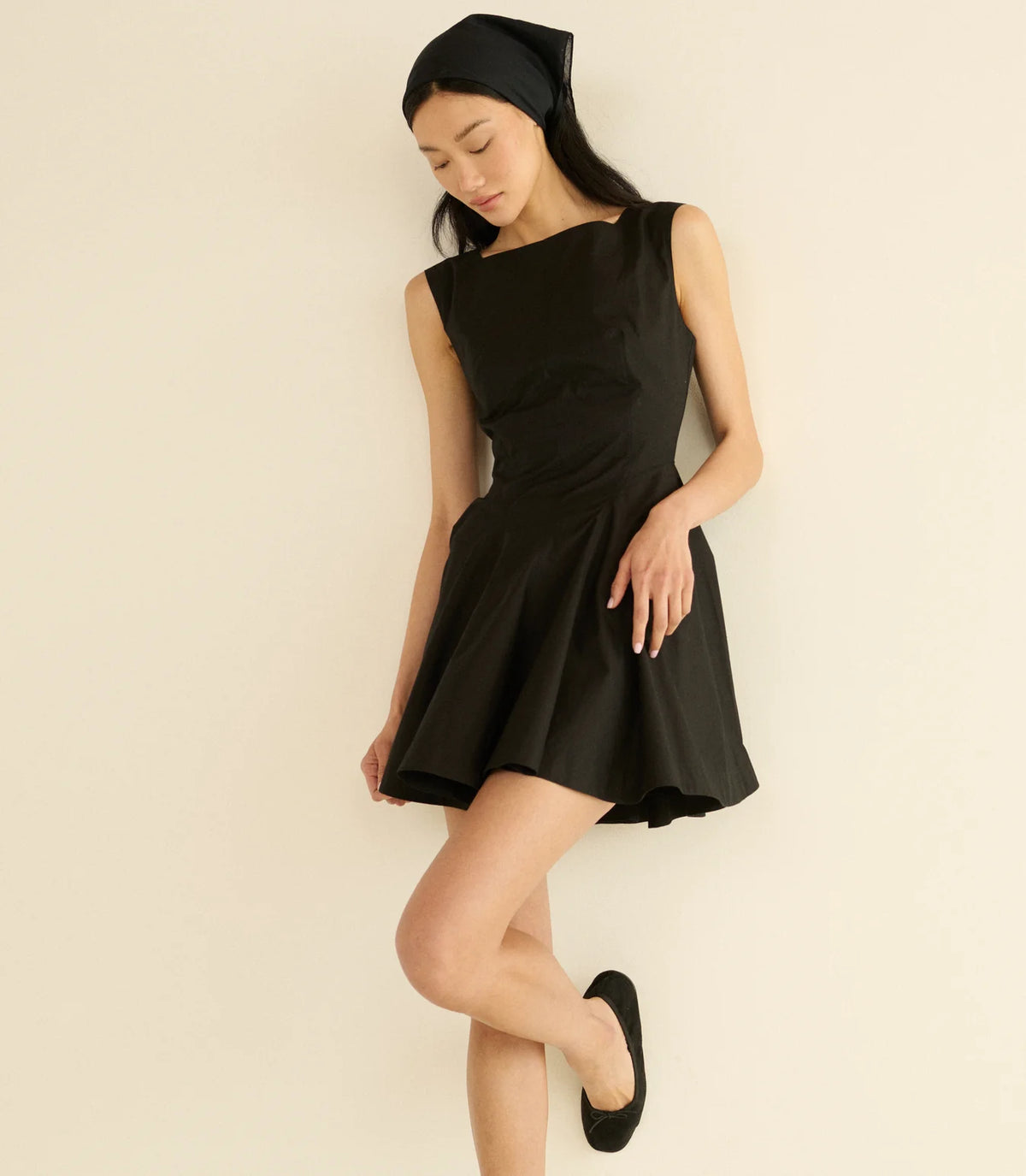 A woman in a sleeveless DOEN Riviette Dress, black flats, and a black headscarf leans gracefully against a light wall, gazing down with one leg slightly bent.