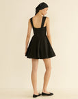 A person stands facing away, wearing a sleeveless black DOEN Riviette Dress with a flared skirt, paired with black ballet flats and a black headscarf, against a plain light background.