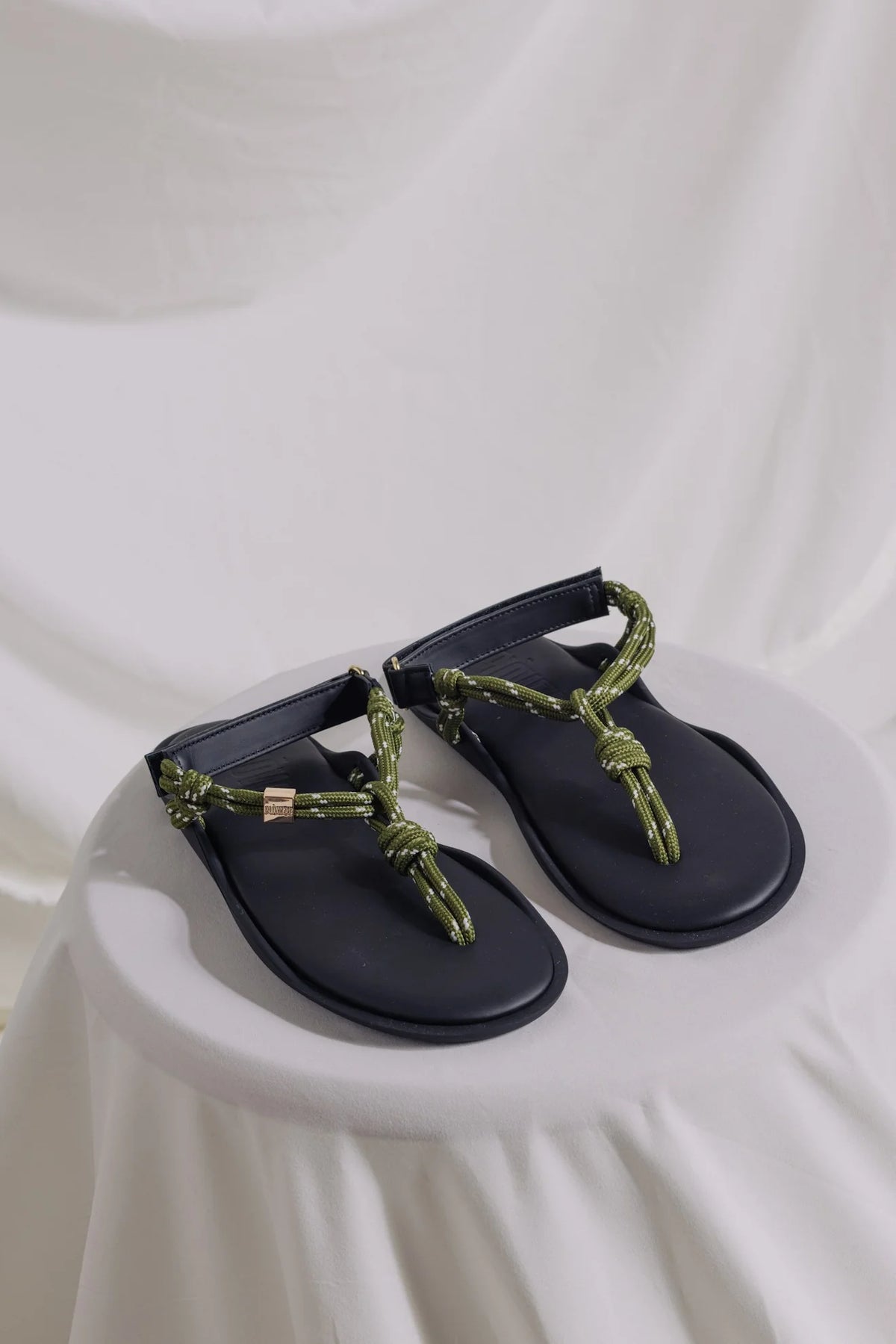 Black adjustable Flowze FLO sandals with green rope straps displayed on a white-draped round surface.