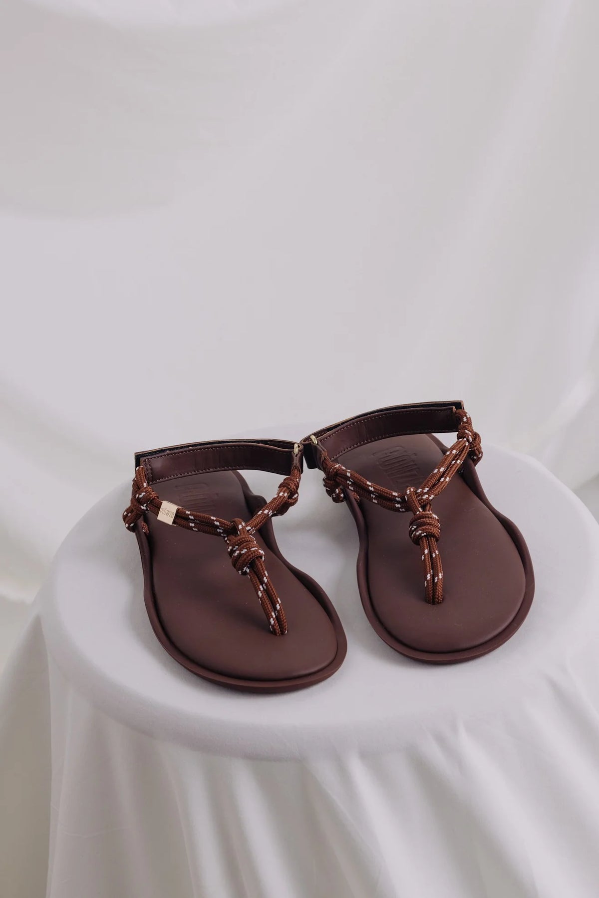 Brown leather Flowze FLO sandals with knotted straps displayed on a white-draped table, Bungalow Furniture.