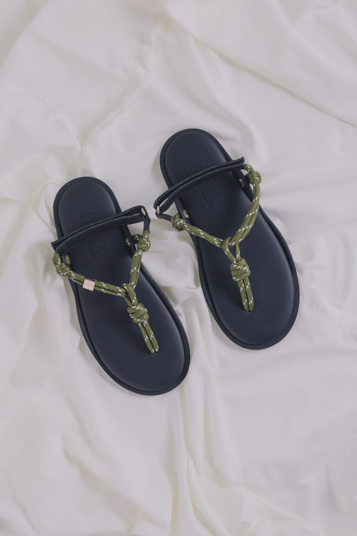 Black leather sandals with green rope straps by Flowze FLO on white fabric background.