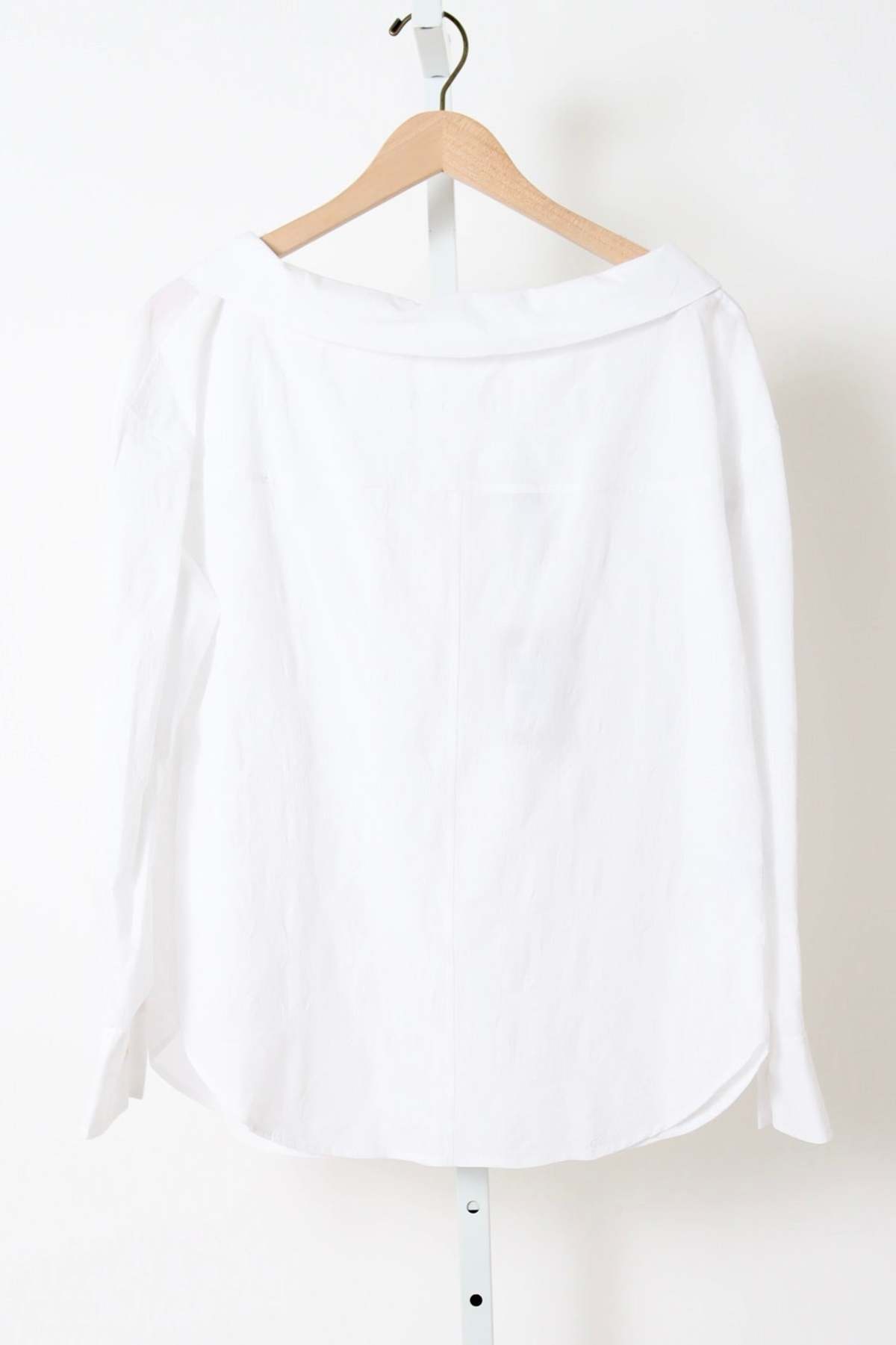 White cotton long-sleeve top by Rachel Comey on wooden hanger against a white wall.