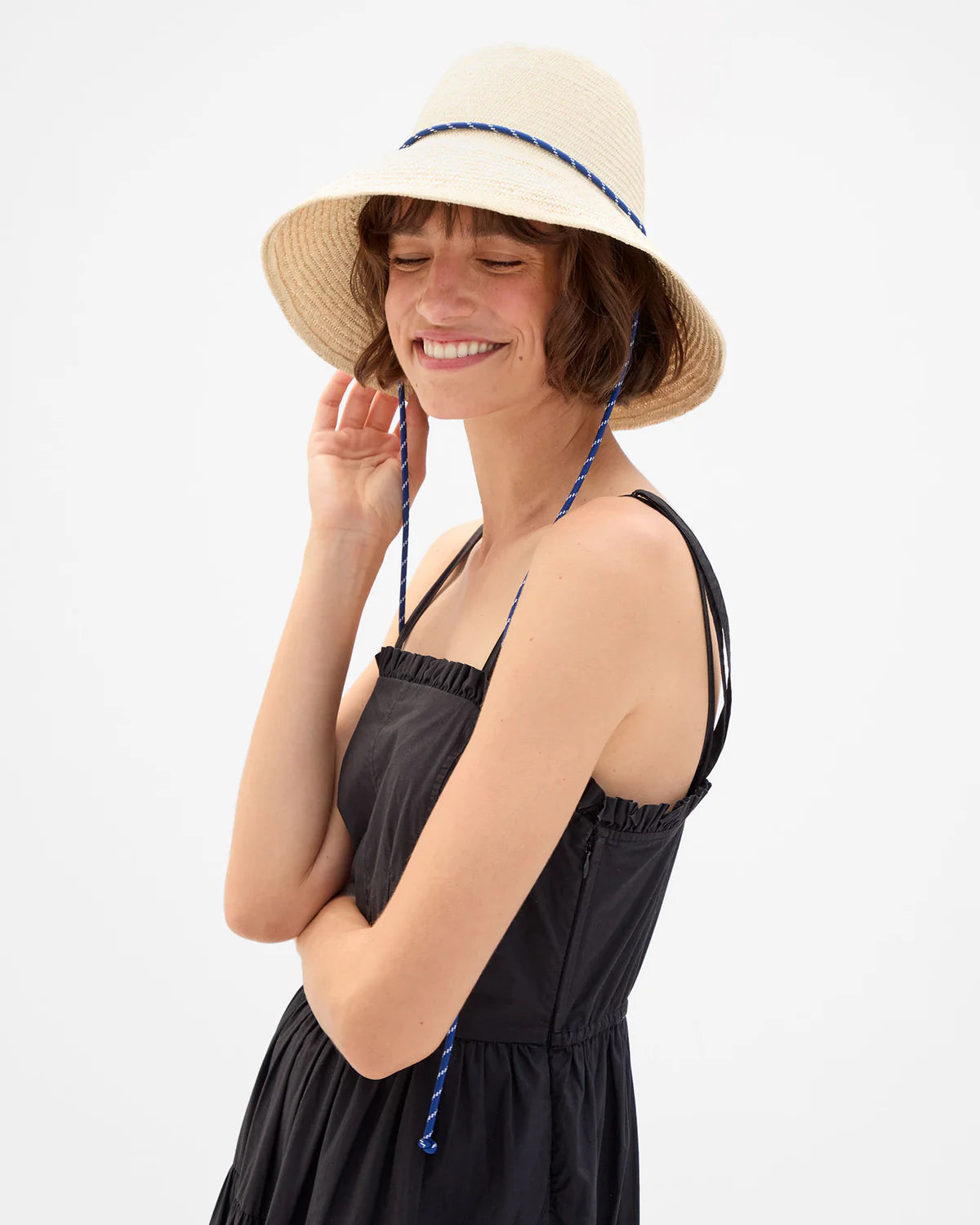 Clare Vivier Romy Hat with sail cord strap on woman in black dress, simple neutral background, stylish look.