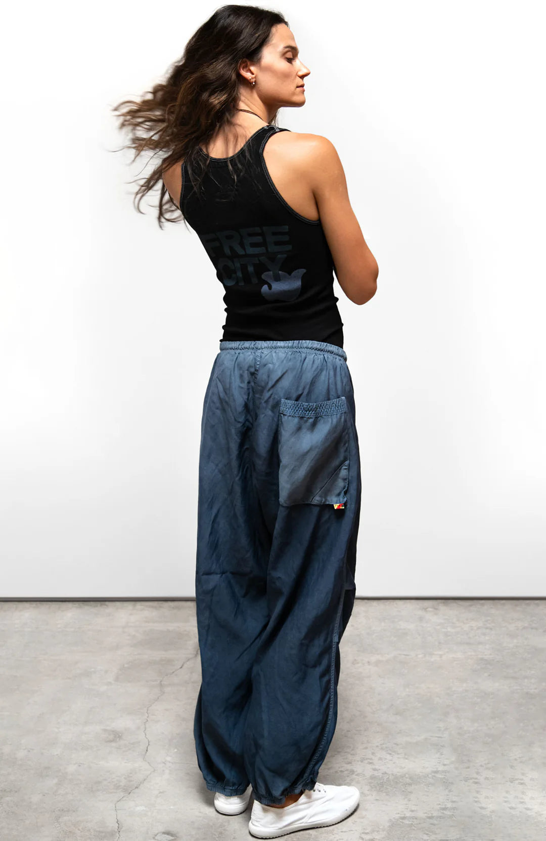 A woman with long brown hair stands on a gray concrete floor facing away, wearing Free City (sparrow, LLC) Matte/Satin Biggy Pant paired with a black sleeveless top. The pants feature a loose fit and large back pocket.