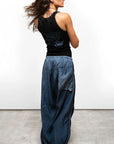 A woman with long brown hair stands on a gray concrete floor facing away, wearing Free City (sparrow, LLC) Matte/Satin Biggy Pant paired with a black sleeveless top. The pants feature a loose fit and large back pocket.