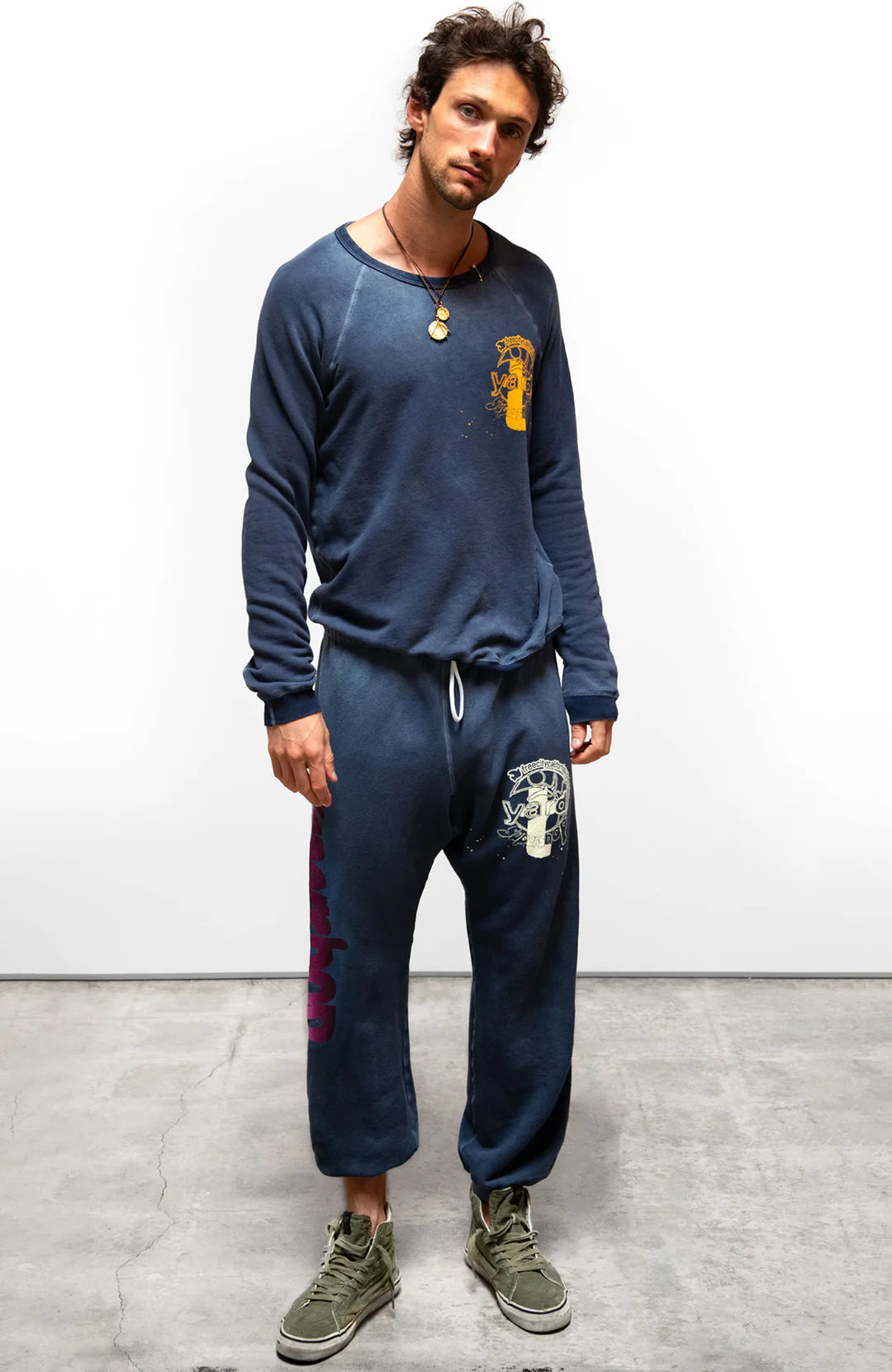 A man stands indoors on a concrete floor, looking at the camera. He wears a navy blue Free City Artyard SUPERSHOP STAMP raglan sweatshirt and matching French terry sweatpants with graphic designs, green sneakers, and a necklace.