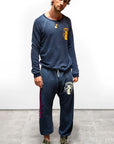 A man stands indoors on a concrete floor, looking at the camera. He wears a navy blue Free City Artyard SUPERSHOP STAMP raglan sweatshirt and matching French terry sweatpants with graphic designs, green sneakers, and a necklace.