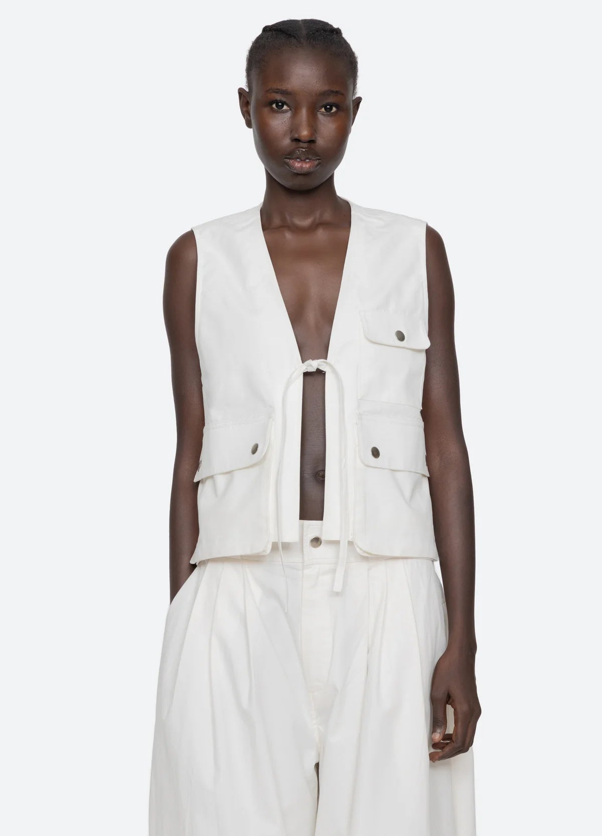 A person faces forward wearing the Sea New York Lois Twill Vest, featuring an oversized utilitarian design with front ties and large pockets, paired with loose white pants against a plain, light-colored background.