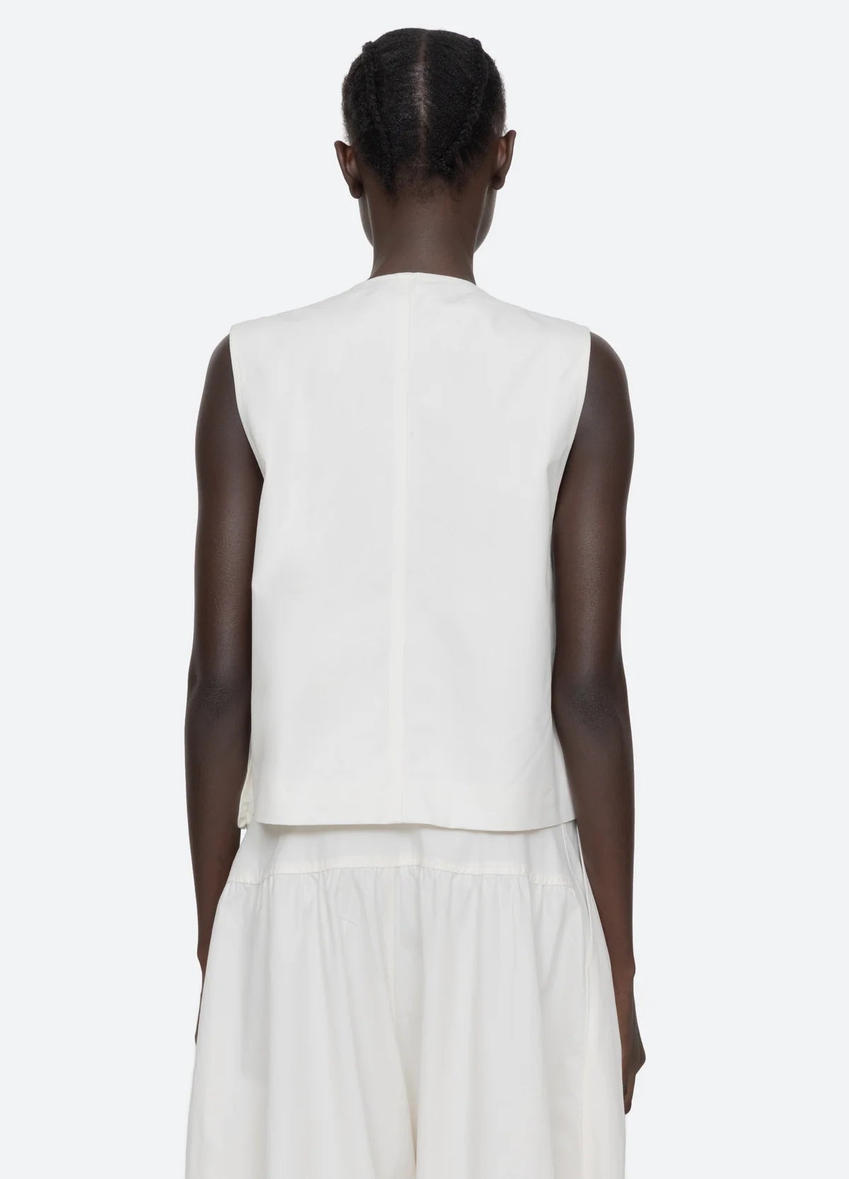 A person with short, braided hair is seen from behind wearing a sleeveless white top, loose white pants, and the Sea New York Lois Twill Vest against a plain light background.