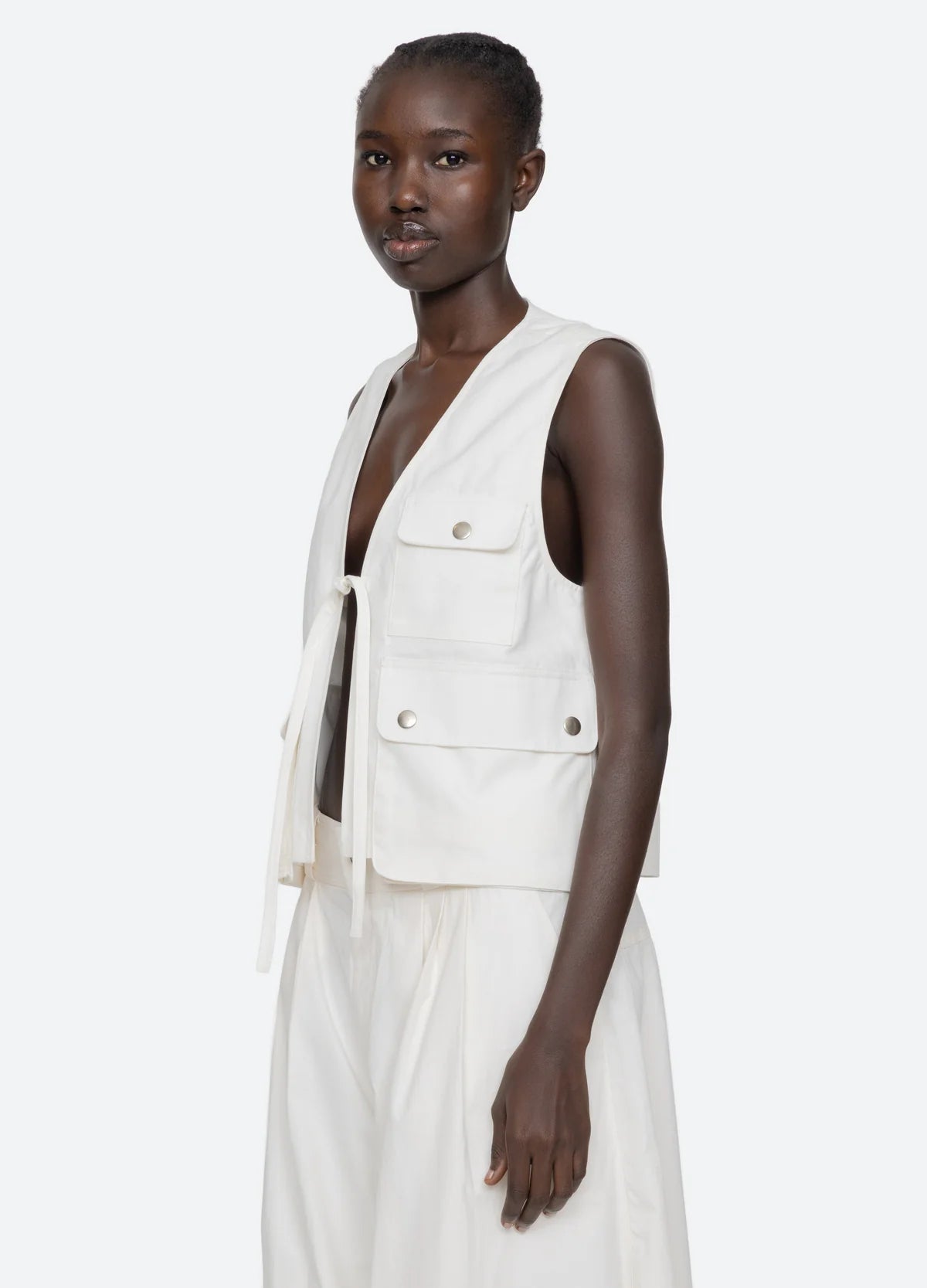 A woman with short hair wears the Lois Twill Vest by Sea New York, an oversized cotton vest with front pockets and ties, paired with a matching white skirt against a plain light background.