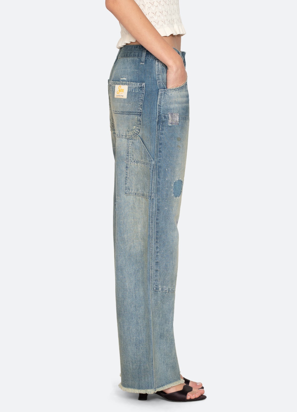 Wearing Sea New York's Claira Mended Jeans—relaxed fit, light blue denim with patchwork and a back pocket label—paired with a white textured top and black sandals, the person stands in side profile.