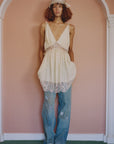 A person with curly hair wears Claira Mended Jeans by Sea New York, a lacy cream dress, sandals, and a decorative headpiece, posing against a pink wall framed by a white arch.