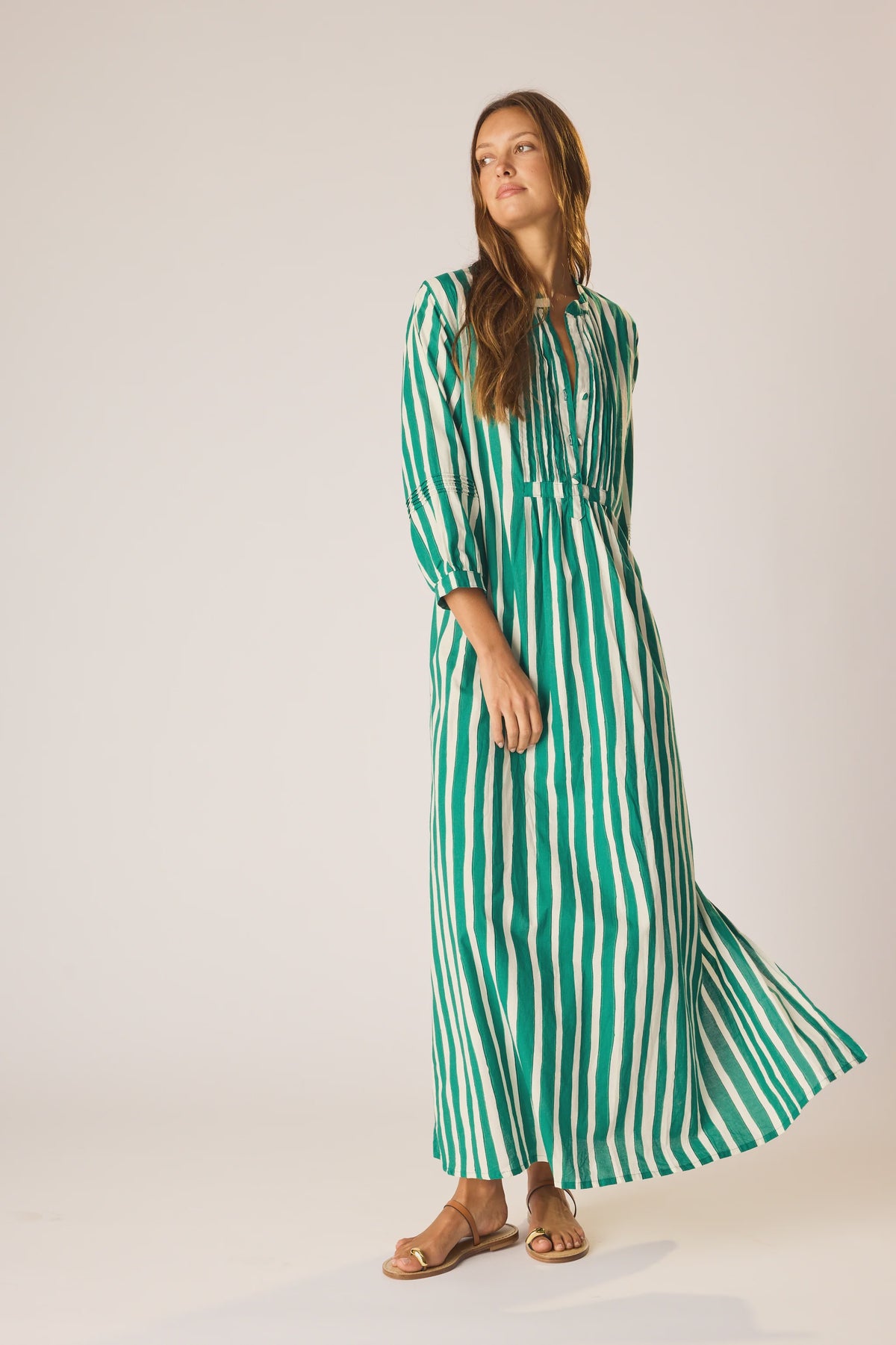 A woman poses in the Sammie Maxi Dress by natalie martin—a green and white vertically striped 100% cotton maxi with three-quarter sleeves and a v-neckline—paired with brown sandals, set against a plain light background.