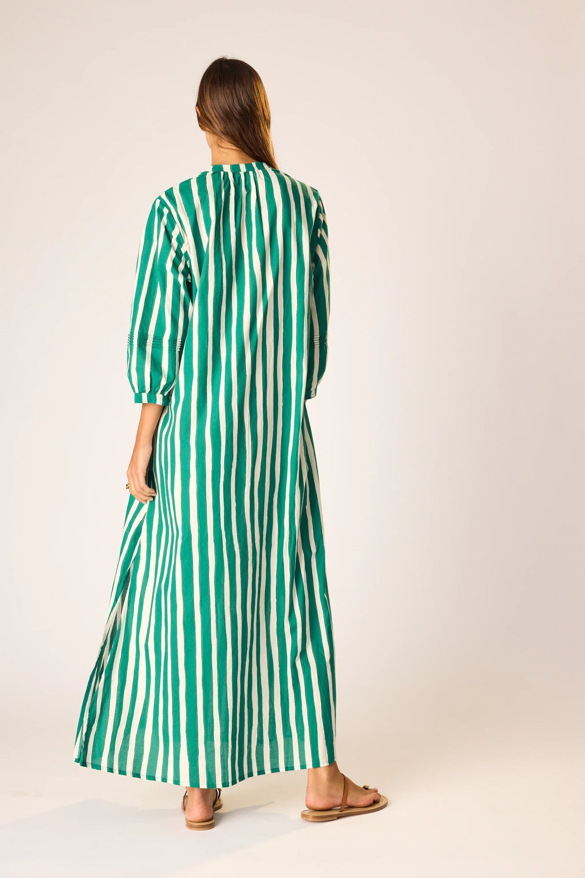 A woman with straight hair stands with her back to the camera in a light studio, wearing the natalie martin Sammie Maxi Dress, a 100% cotton dress featuring vertical green and white stripes and paired with flat sandals.