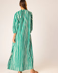 A woman with straight hair stands with her back to the camera in a light studio, wearing the natalie martin Sammie Maxi Dress, a 100% cotton dress featuring vertical green and white stripes and paired with flat sandals.