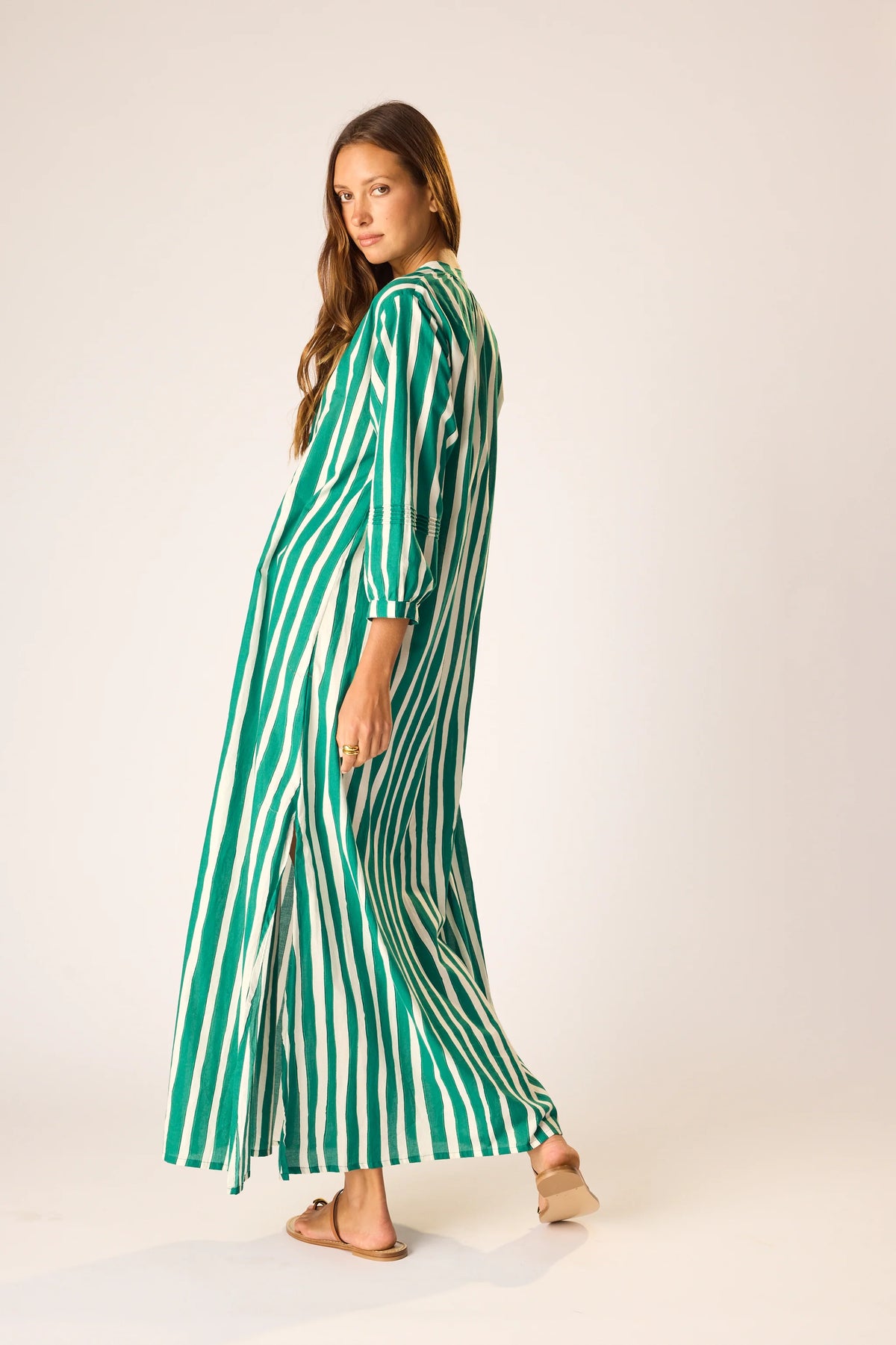 A woman in the natalie martin Sammie Maxi Dress—a long, green and white vertically striped v-neck dress—stands barefoot against a light background, glancing over her shoulder with a relaxed look.