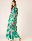 A woman in the natalie martin Sammie Maxi Dress—a long, green and white vertically striped v-neck dress—stands barefoot against a light background, glancing over her shoulder with a relaxed look.