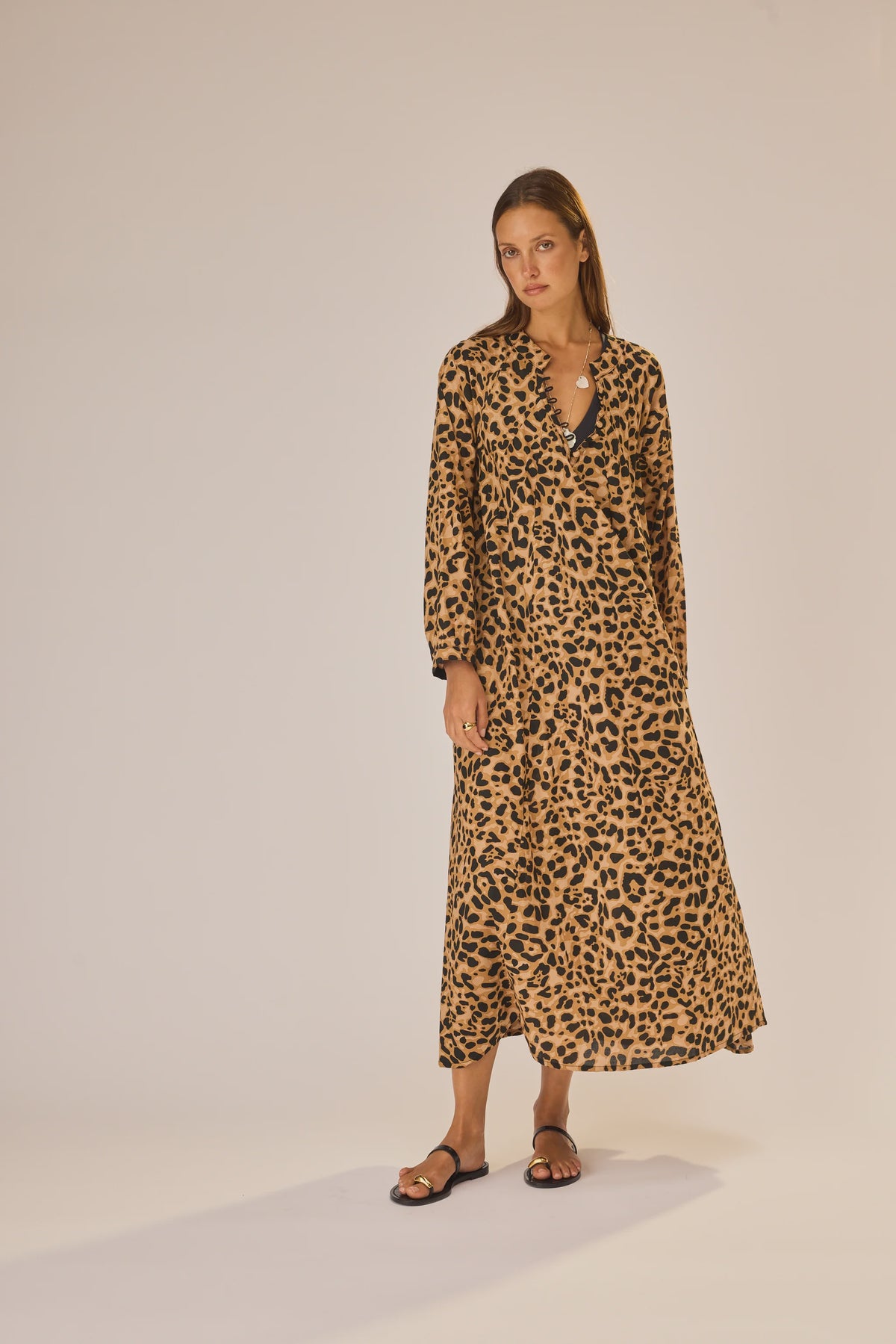 A woman with straight hair and a neutral expression wears the natalie martin Fiore Maxi, a long, loose-fitting A-line leopard print dress with long sleeves, paired with black sandals against a plain background.