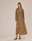 A woman with straight hair and a neutral expression wears the natalie martin Fiore Maxi, a long, loose-fitting A-line leopard print dress with long sleeves, paired with black sandals against a plain background.