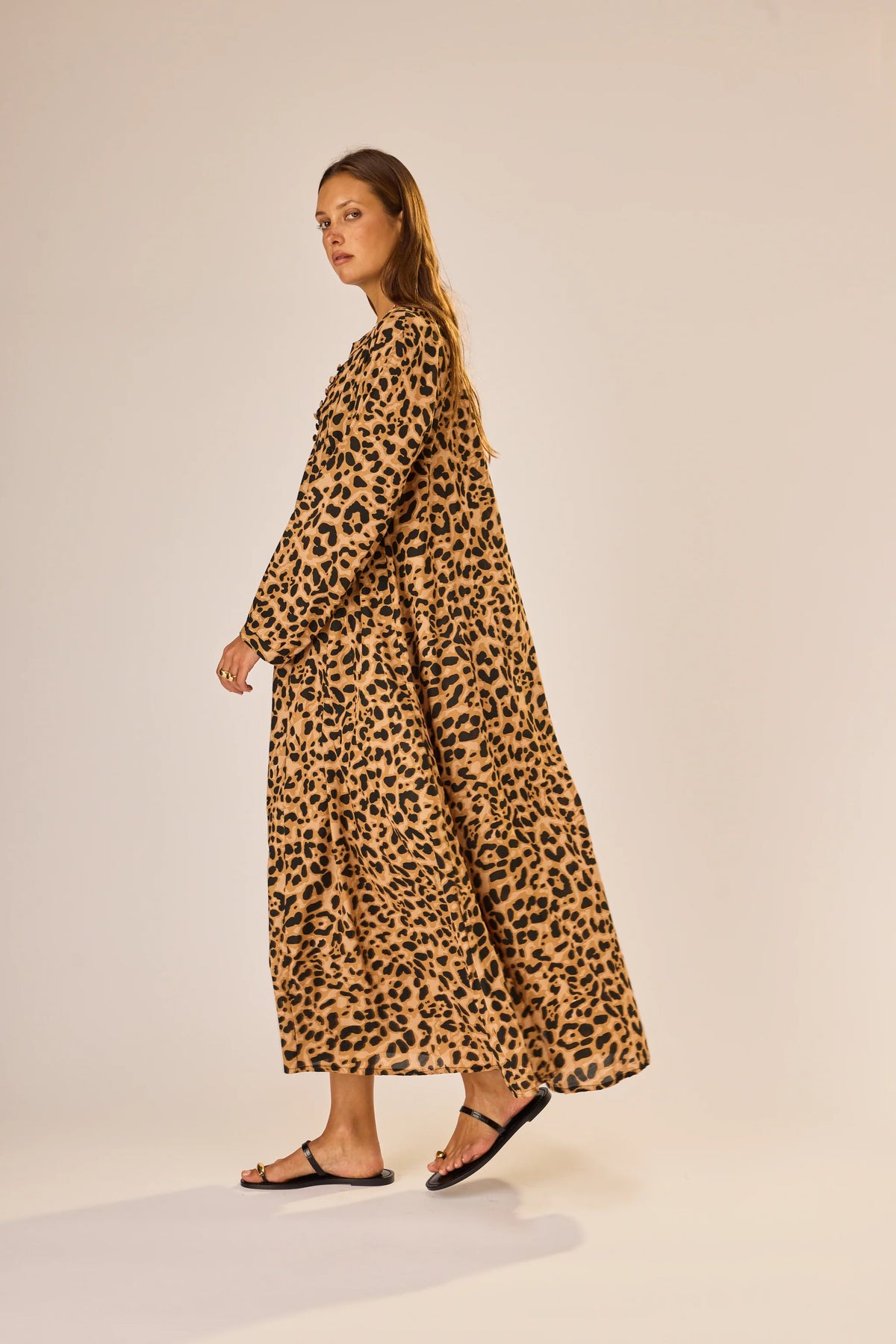 A woman stands sideways in the Fiore Maxi by natalie martin—a long, loose-fitting leopard print A-line dress with long sleeves. She has long hair, wears black sandals, and poses against a plain light-colored background.