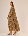 A woman stands sideways in the Fiore Maxi by natalie martin—a long, loose-fitting leopard print A-line dress with long sleeves. She has long hair, wears black sandals, and poses against a plain light-colored background.