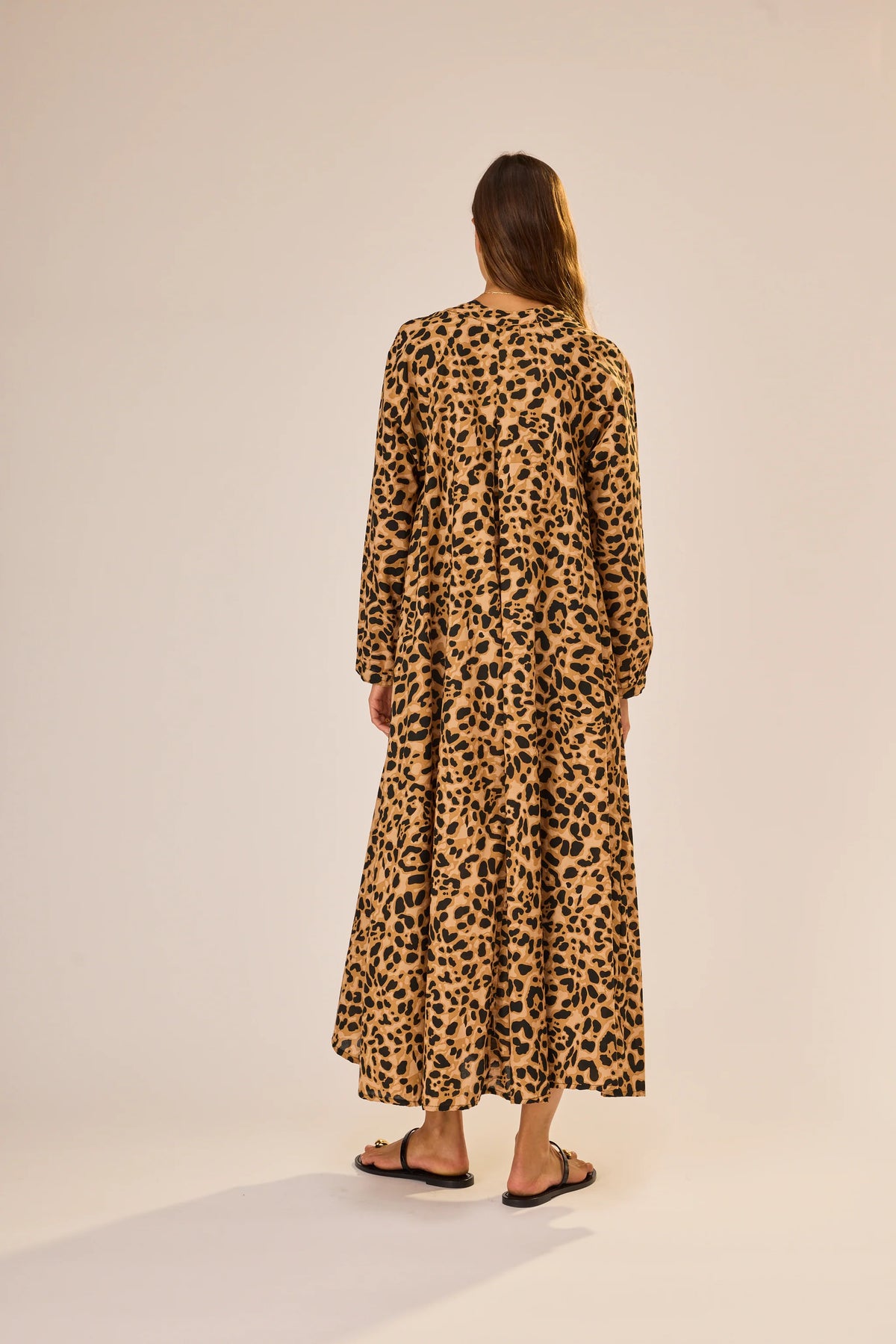 A woman with long brown hair wears the natalie martin Fiore Maxi, an ankle-length, long sleeve A-line dress in tan and black leopard print, paired with flat sandals. She stands facing away against a neutral background.